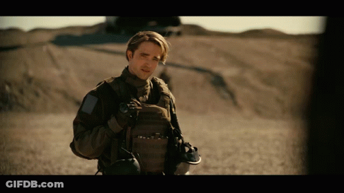A Man In A Military Uniform Stands In A Desert Holding A Helmet GIF