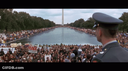 A Man In A Military Uniform Stands In Front Of A Crowd Holding Signs Including One That Says " Peace " GIF