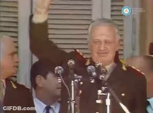 A Man In A Military Uniform Stands In Front Of Two Microphones And Waves His Hand GIF