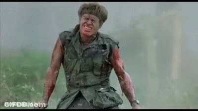 A Man In A Military Uniform With Blood On His Face And Arms Is Standing In A Field . GIF