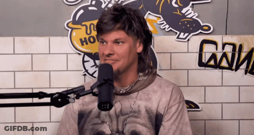A Man In A Mullet Stands In Front Of A Microphone In Front Of A Sticker That Says Ho GIF