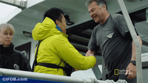 A Man In A National Geographic Shirt Shakes Hands With Another Man On A Boat GIF