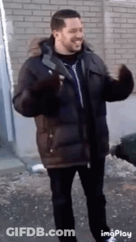 A Man In A Parka Is Standing In Front Of A Brick Wall And Waving . GIF