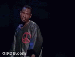 A Man In A Peace Sign Shirt Is Standing In Front Of A Microphone With The Words Next Time Above Him GIF