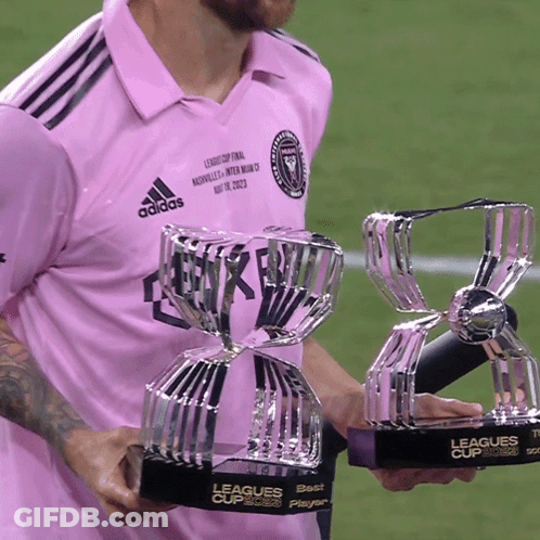 A Man In A Pink Adidas Shirt Is Holding Two League Cup Trophies GIF