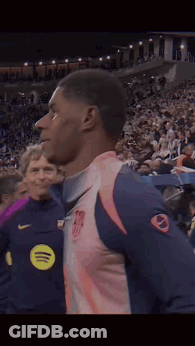 A Man In A Pink And Blue Jersey Is Standing In Front Of A Crowd Of People . GIF