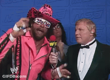 A Man In A Pink Jacket Is Talking Into A Microphone While Another Man In A Tuxedo Watches . GIF