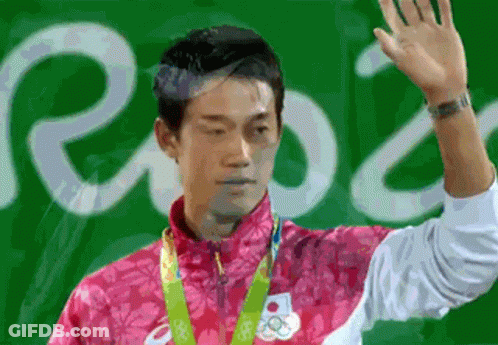 A Man In A Pink Jacket With A Medal Around His Neck Is Waving His Hand In Front Of A Sign That Says Rio GIF
