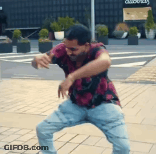 A Man In A Pink Shirt And Blue Jeans Is Dancing On A Brick Sidewalk . GIF
