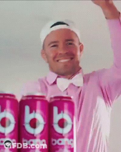 A Man In A Pink Shirt And Bow Tie Is Standing Next To A Row Of Cans Of Bang GIF