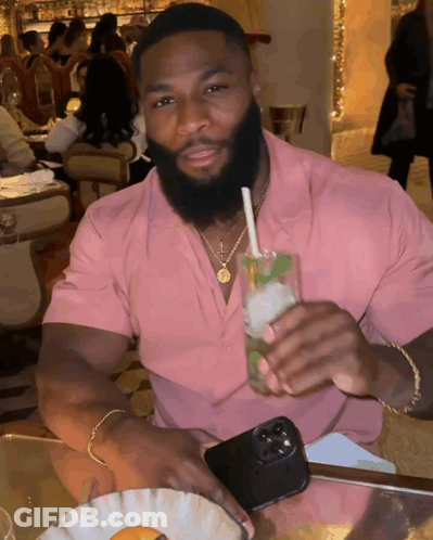 A Man In A Pink Shirt Is Sitting At A Table Holding A Drink GIF