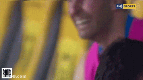 A Man In A Pink Shirt Is Smiling In Front Of A Tico Sports Sign GIF