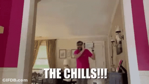 A Man In A Pink Shirt Is Standing In A Living Room With The Words The Chills Written On The Wall Behind Him . GIF