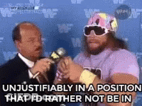 A Man In A Pink Shirt Is Talking Into A Microphone While A Man In A Suit Looks On . GIF