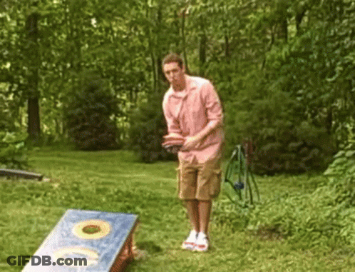 A Man In A Pink Shirt Is Throwing A Frisbee In A Cornhole Board GIF