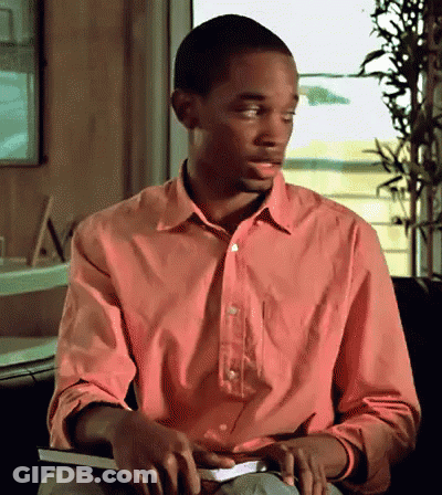 A Man In A Pink Shirt Sits In A Chair Holding A Book GIF