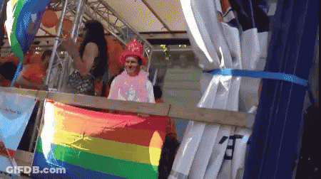A Man In A Pink Shirt Stands Behind A Rainbow Flag With Chinese Characters On It GIF