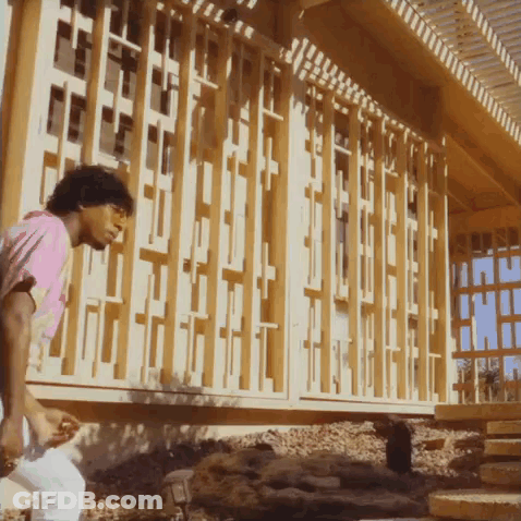 A Man In A Pink Shirt Stands In Front Of A Building With A Wooden Fence GIF