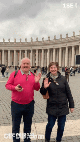 A Man In A Pink Sweater And A Woman In A Black Coat Are Standing In Front Of A Building With The Date Jan 06 2023 GIF