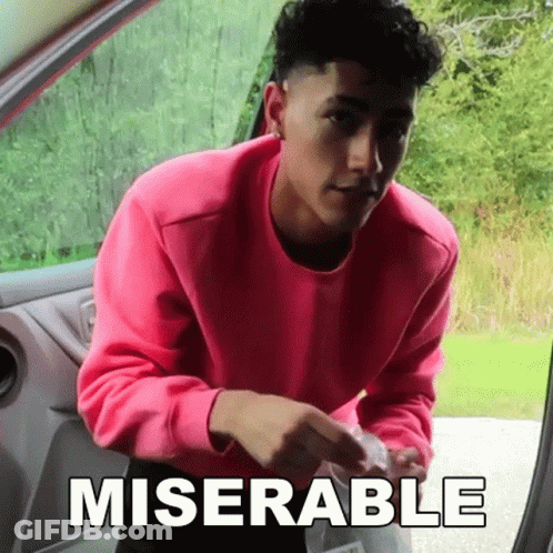 A Man In A Pink Sweater Is Standing In The Doorway Of A Car With The Word Miserable Written On The Side . GIF