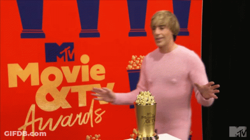 A Man In A Pink Sweater Stands In Front Of A Movie And TV Awards Sign GIF