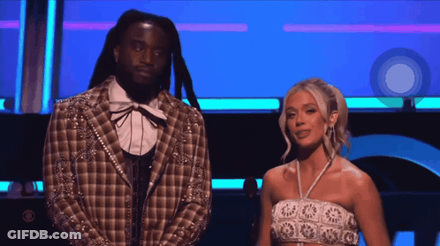 A Man In A Plaid Jacket Stands Next To A Woman In A White Crop Top GIF