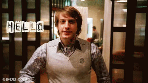 A Man In A Plaid Shirt And A White Vest Says Hello GIF