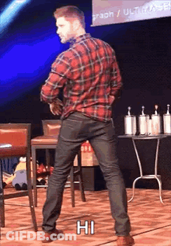 A Man In A Plaid Shirt And Black Jeans Is Standing On A Stage With His Hands In His Pockets . GIF