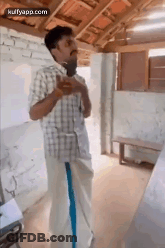 A Man In A Plaid Shirt And Blue Pants Is Standing In A Room With A Microphone In His Hand . GIF