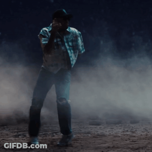 A Man In A Plaid Shirt And Cowboy Hat Is Standing In A Dark Room GIF