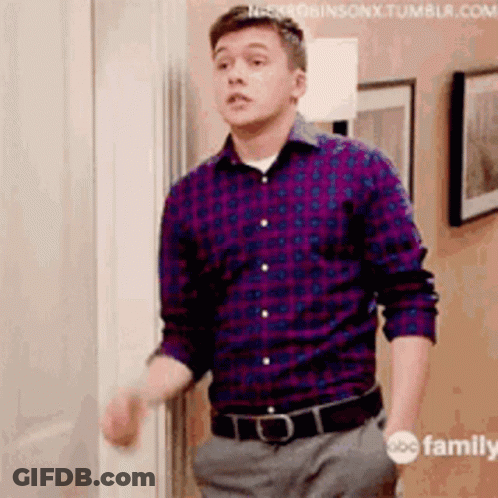 A Man In A Plaid Shirt And Gray Pants Is Standing In Front Of A Door . GIF