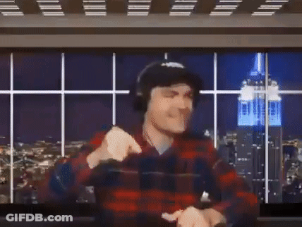 A Man In A Plaid Shirt And Headphones Is Raising His Arms In The Air In Front Of A Large Screen . GIF