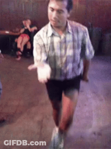 A Man In A Plaid Shirt And Shorts Is Dancing In A Room With People In The Background . GIF