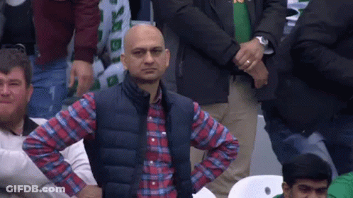 A Man In A Plaid Shirt And Vest Is Sitting In A Crowd Of People With His Hands On His Hips . GIF