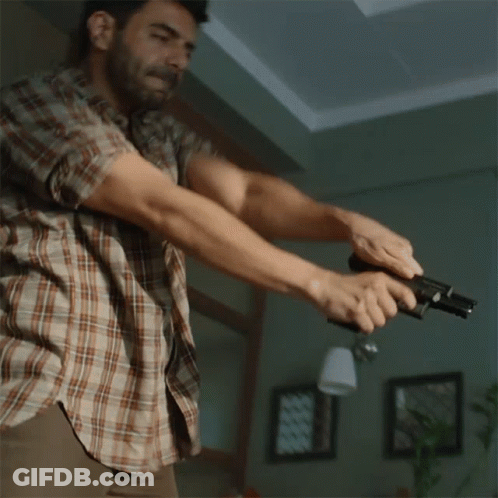 A Man In A Plaid Shirt Holds A Gun In His Hand GIF