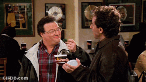 A Man In A Plaid Shirt Is Feeding Another Man Something GIF