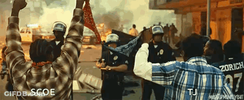 A Man In A Plaid Shirt Is Holding A Bandana In Front Of Riot Police . GIF
