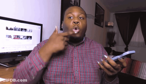 A Man In A Plaid Shirt Is Holding A Cell Phone In Front Of A Computer Monitor GIF