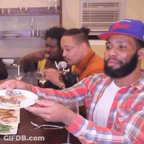 A Man In A Plaid Shirt Is Holding A Plate Of Food In Front Of A Group Of Men GIF