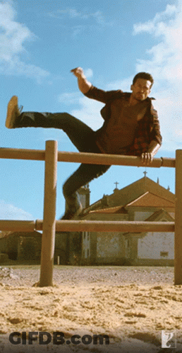 A Man In A Plaid Shirt Is Jumping Over A Wooden Railing GIF