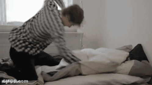 A Man In A Plaid Shirt Is Kneeling On A Bed With A Pillow . GIF