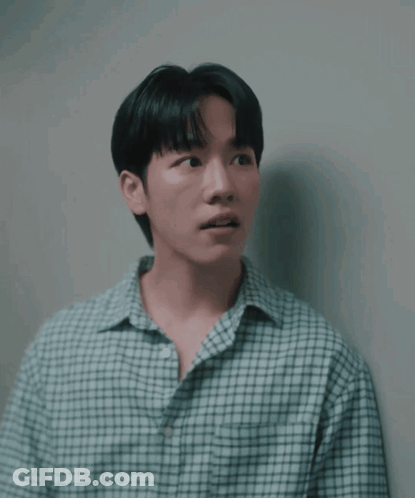 A Man In A Plaid Shirt Is Leaning Against A White Wall GIF