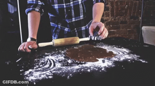 A Man In A Plaid Shirt Is Rolling Out A Piece Of Dough GIF