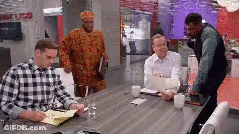 A Man In A Plaid Shirt Is Sitting At A Conference Table With Other Men GIF