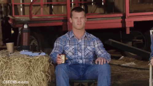 A Man In A Plaid Shirt Is Sitting In Front Of A Bale Of Hay GIF