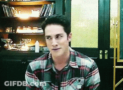 A Man In A Plaid Shirt Is Sitting In Front Of A Bookcase GIF