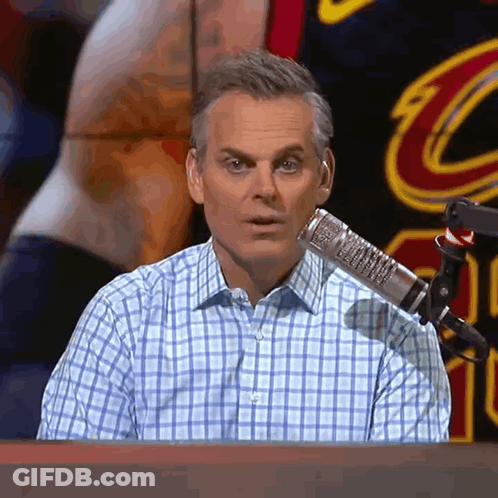 A Man In A Plaid Shirt Is Sitting In Front Of A Microphone With A Cleveland Cavaliers Jersey In The Background GIF