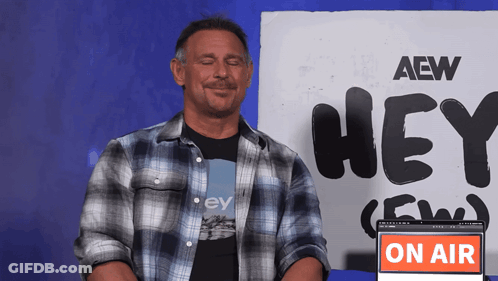 A Man In A Plaid Shirt Is Sitting In Front Of A Sign That Says Hey GIF
