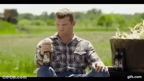A Man In A Plaid Shirt Is Sitting On A Truck In A Field Holding A Glass Of Beer . GIF