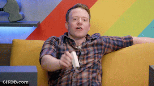 A Man In A Plaid Shirt Is Sitting On A Yellow Couch Holding A White Controller GIF
